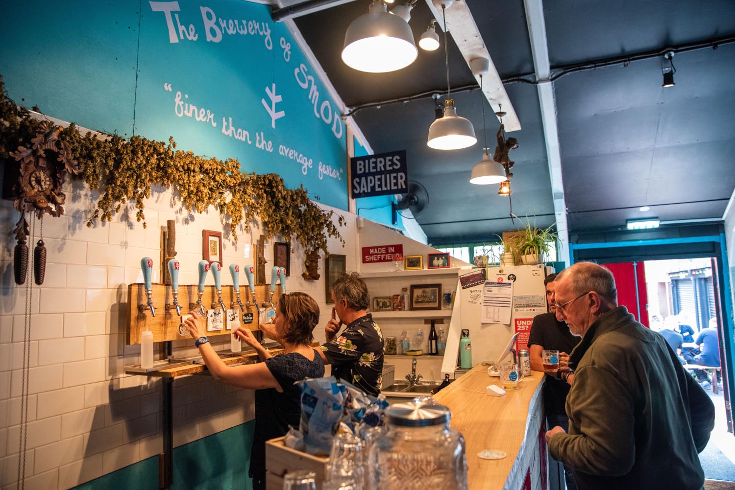 Two people are behind a bar, pulling pints of beer, as two customers wait on the other side.