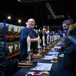 A long bar, at a beer festival, with many beer pumps. A man behind the bar is serving two people.