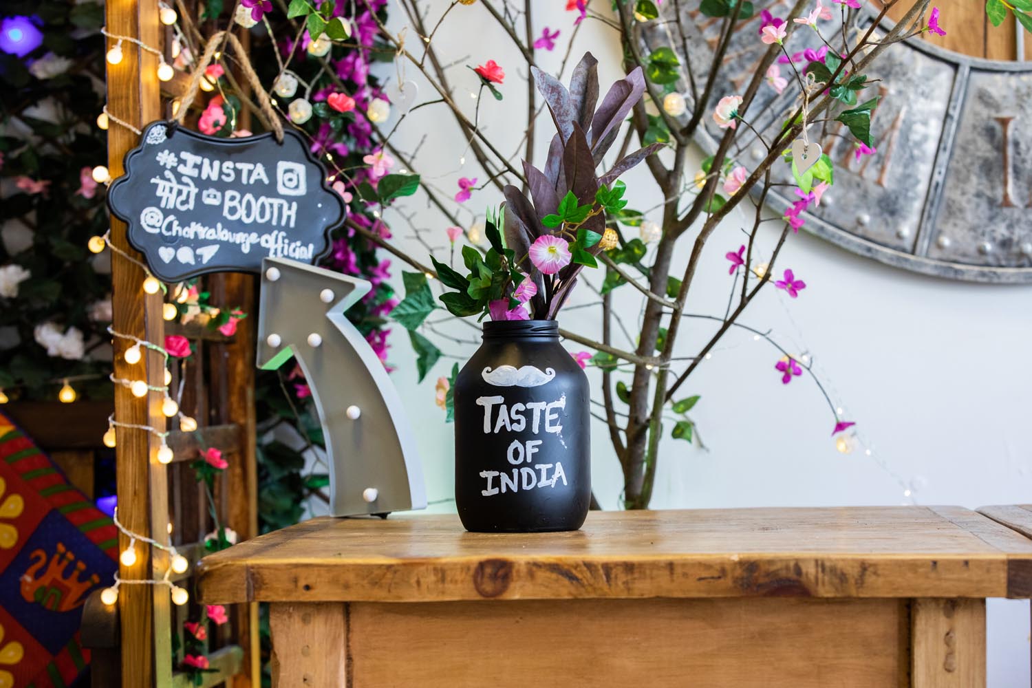 A small wooden table with a floral display and a bottle, which has the words 'Taste of India' painted on it.