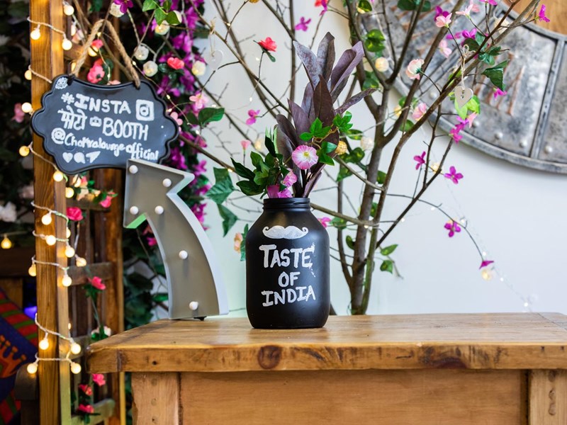 A small wooden table with a floral display and a bottle, which has the words 'Taste of India' painted on it.