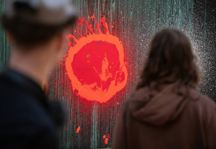 Two people stand observing a piece of abstract art. The artwork features a vivid red splatter resembling an explosion or sunburst, set against a dark, textured background with streaks of green and white. The red element is the focal point, creating a striking contrast and dynamic visual impact.