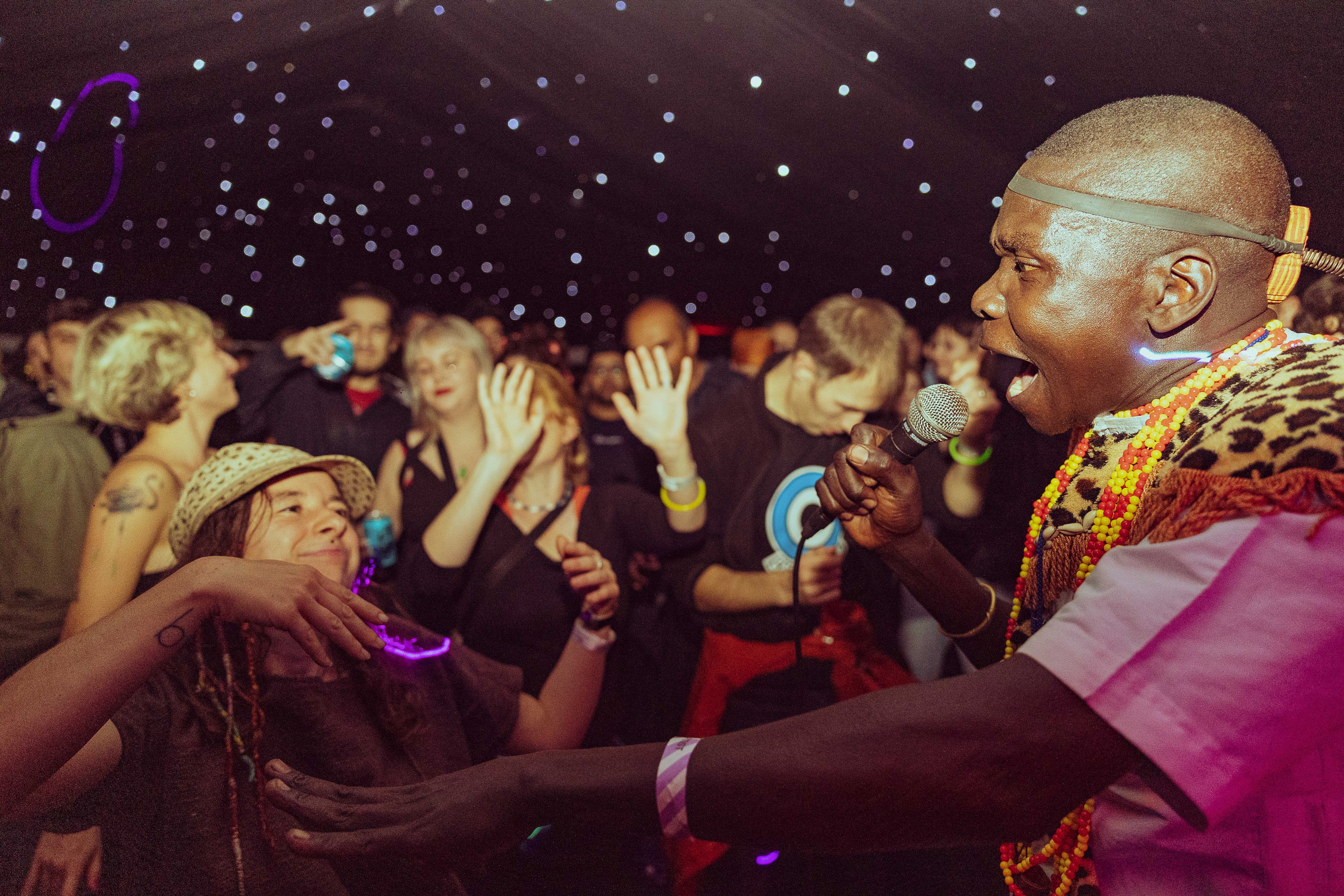 A performer holding a microphone interacts with the crowd under a dark ceiling filled with small glowing lights, resembling stars. The audience is gathered closely, some wearing glow accessories, and the atmosphere suggests a lively music or dance event.