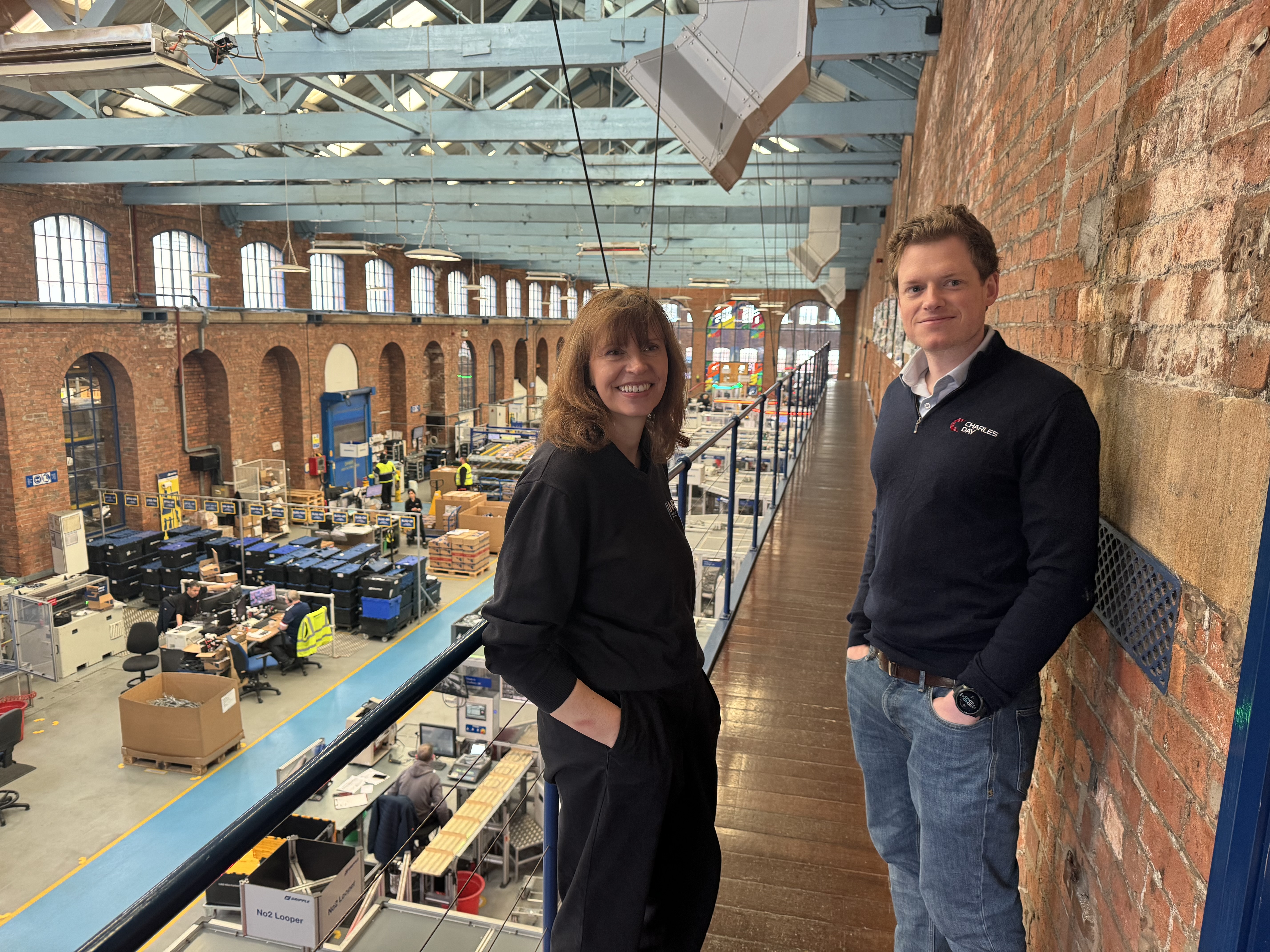 Two people standing on an upper-level walkway inside a large industrial building with exposed brick walls and blue-painted steel beams. The walkway overlooks a spacious factory floor filled with workstations, machinery, and shelving units. The area below is organized with tables, equipment, and workers in high-visibility clothing. Large arched windows line the brick walls, allowing natural light into the space.