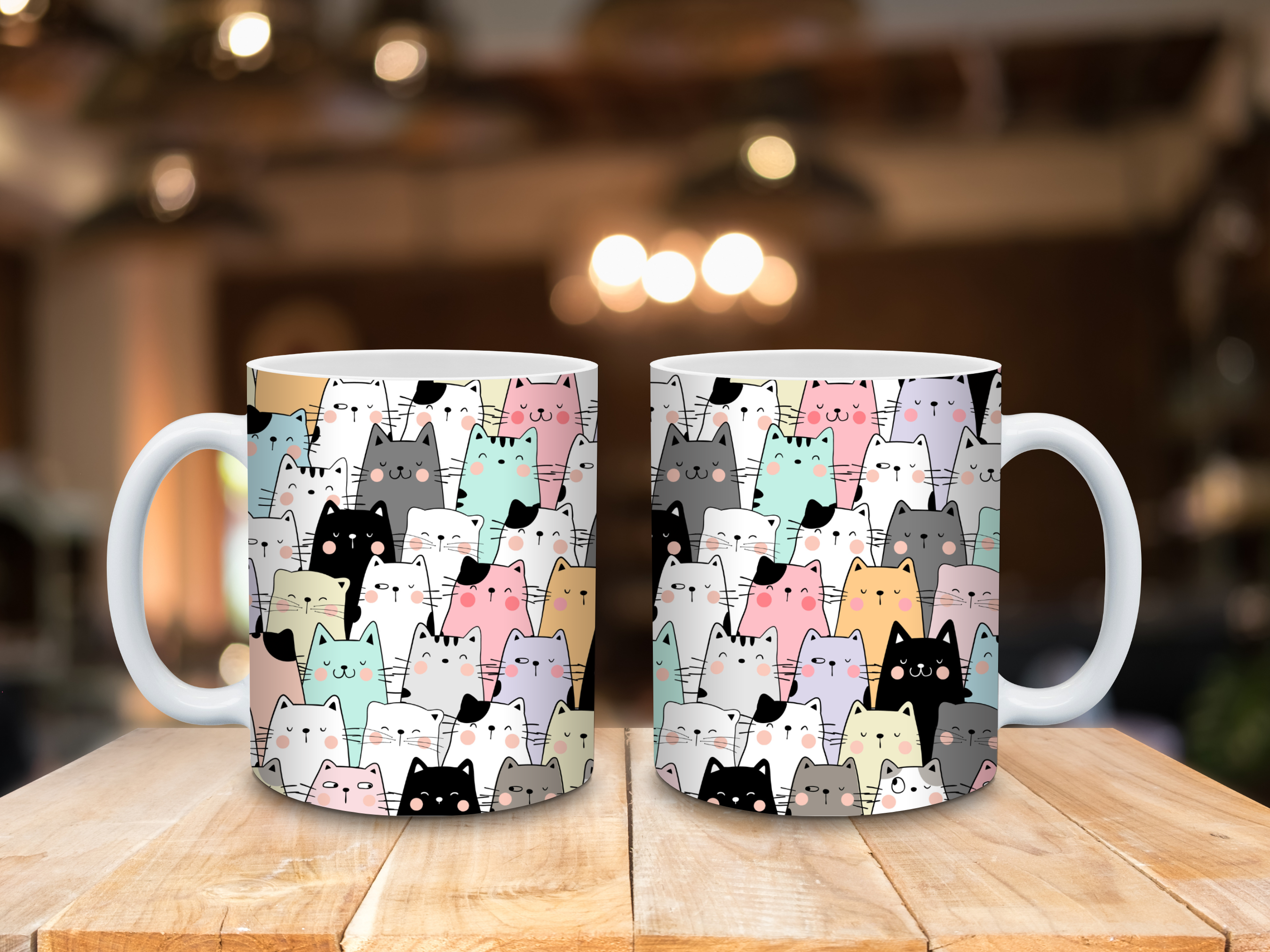 Two ceramic mugs placed on a wooden surface, each featuring a playful pattern of cartoon-style cats in pastel colors including pink, mint green, yellow, gray, and black. The background shows a softly blurred indoor setting with warm lighting.