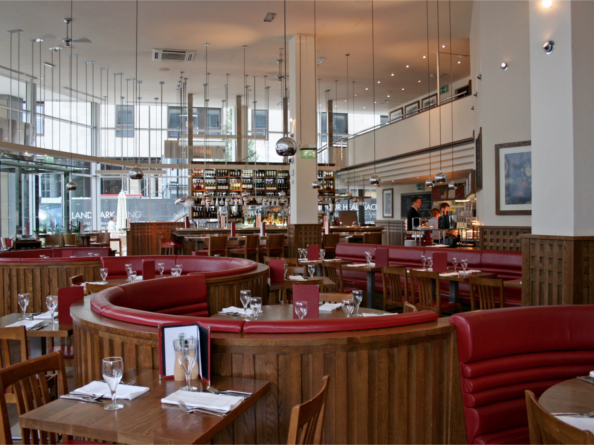 The interior at Piccolino. Bright, modern restaurant interior with large windows, high ceilings, and hanging pendant lights. The space features curved wooden booths with red leather seating and neatly set tables with glassware and menus. A long bar at the back is stocked with bottles, and the overall design combines warm wood tones with contemporary elements.