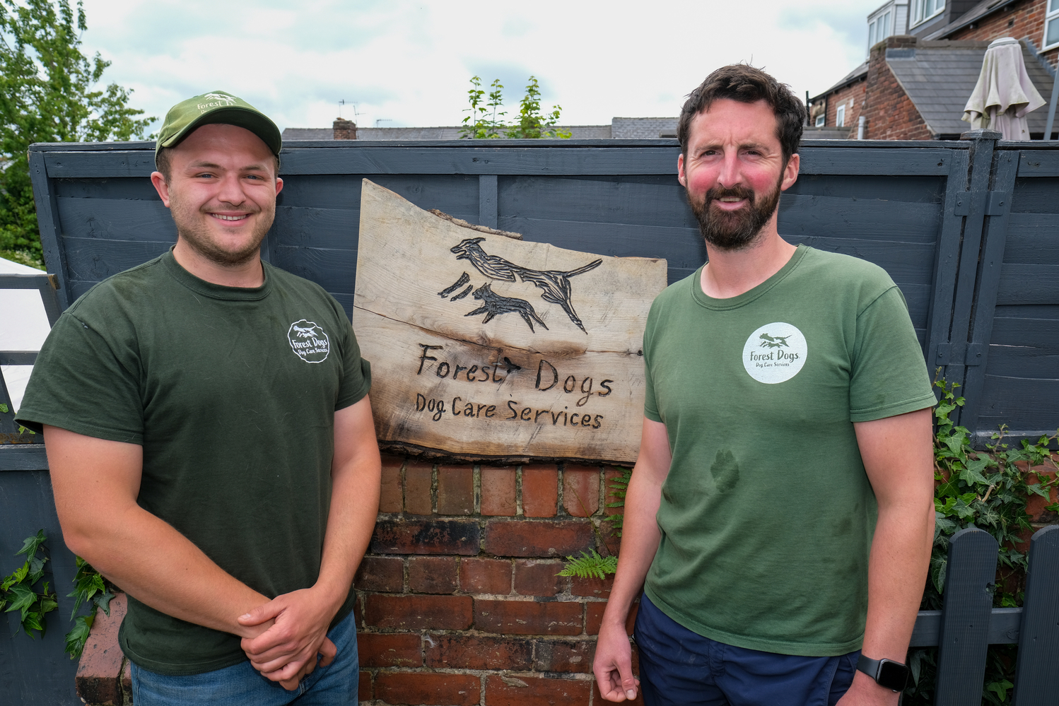 Two men are stood next to a fence that has a sign on itthat reads 'Forrest Dogs'.