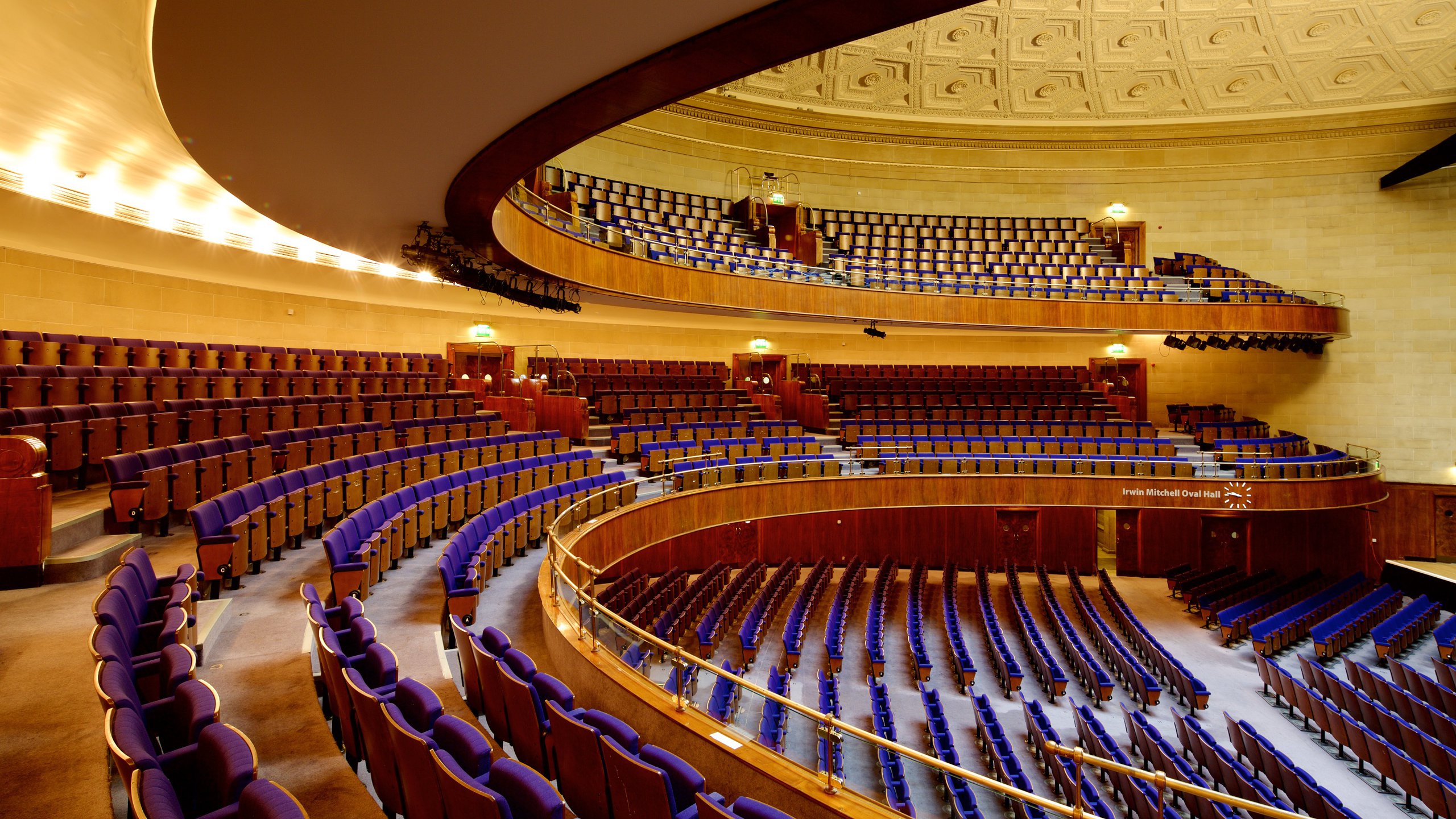 An elegant auditorium with tiered seating arranged in a semi-circular layout. The seats are upholstered in deep blue fabric, and the space features multiple balconies with matching seating. Warm lighting illuminates the curved wooden panels and the ornate, patterned ceiling. The overall design conveys a grand and formal atmosphere suitable for concerts, lectures, or large events.