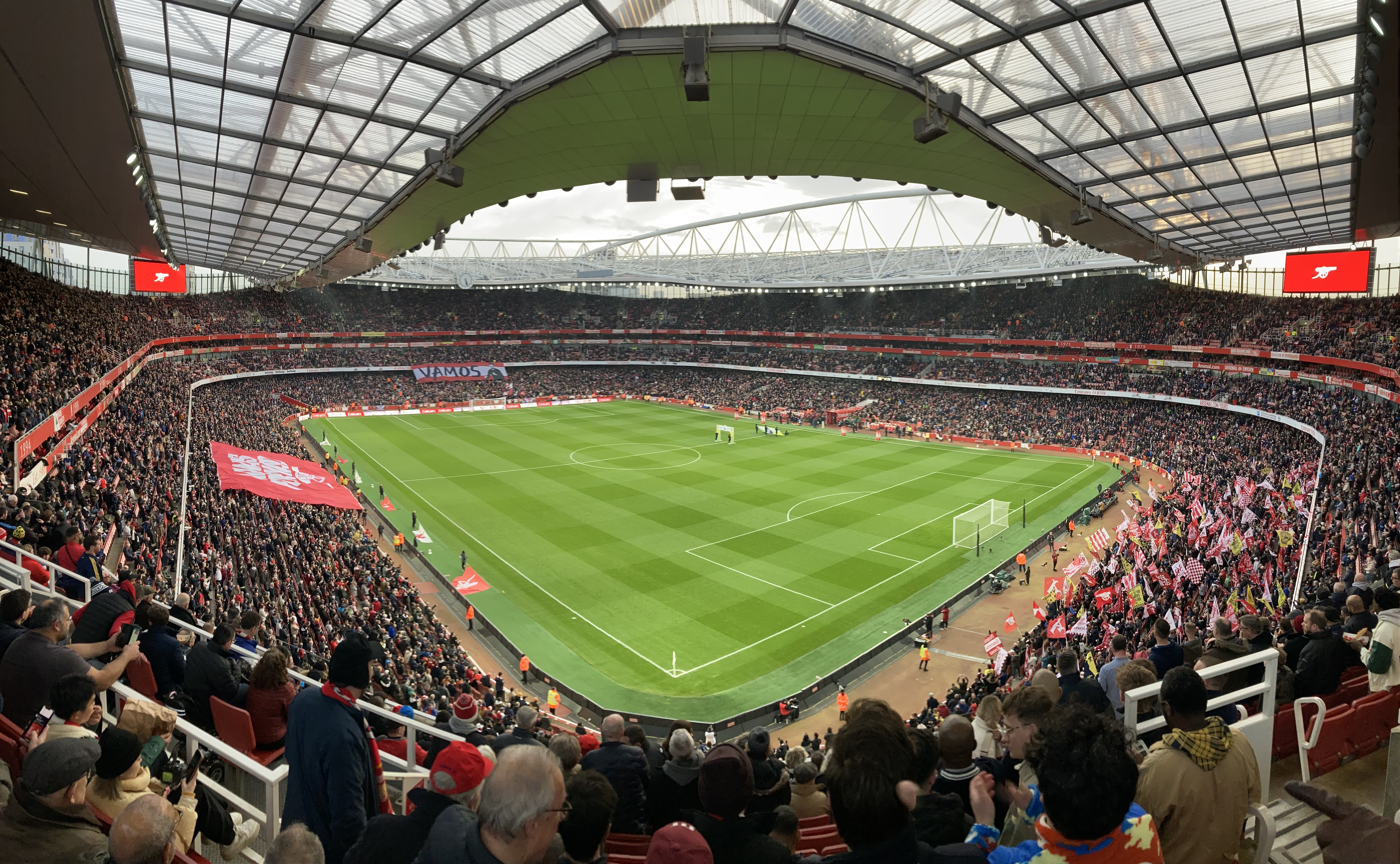 Panoramic view of a packed football stadium with fans filling the stands. The green pitch is set for a match, with goalposts visible at both ends. Large red banners and flags are displayed by supporters, and the stadium roof arches overhead with a modern design.