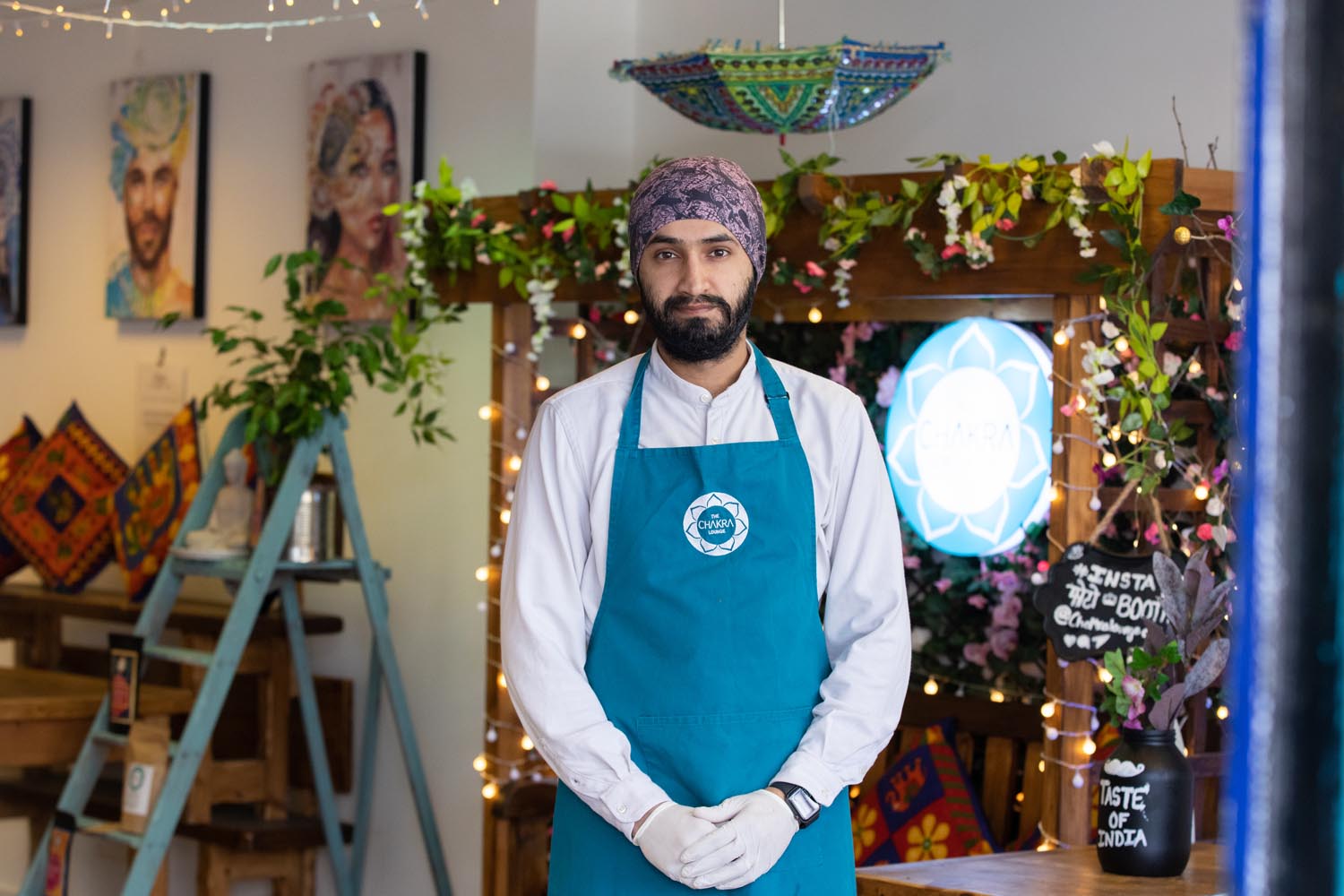 Person wearing a teal apron and white gloves stands inside a decorated shop or café. The background features hanging string lights, colorful cushions, framed artwork, and a wooden structure adorned with flowers and greenery. A round illuminated sign and a chalkboard with text are also visible.