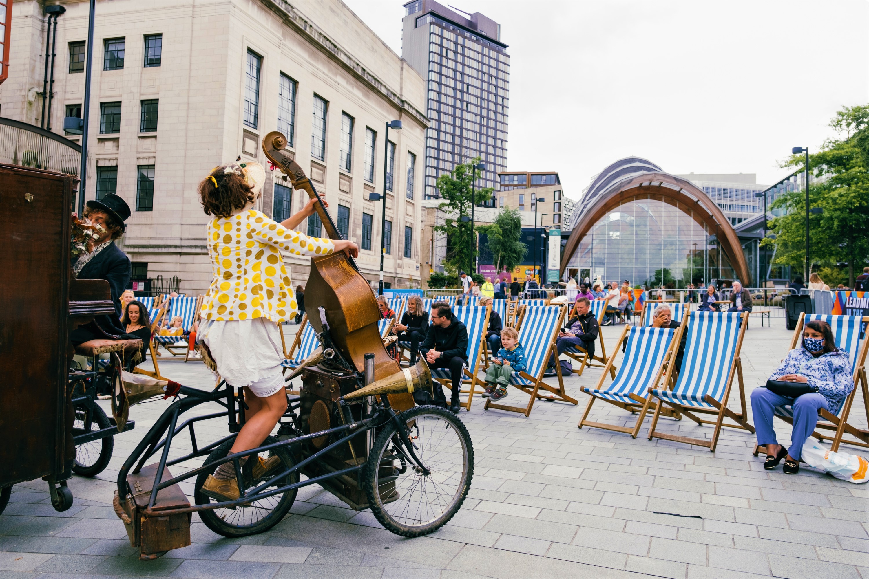 People sat in deckchairs are watching street performers as part of Together In The Square Outdoor Street Theatre.