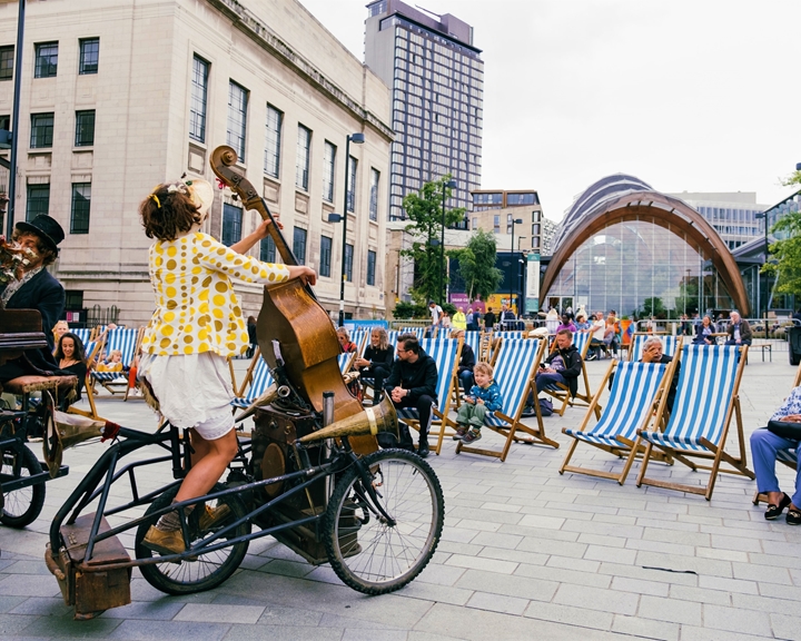 People sat in deckchairs are watching street performers as part of Together In The Square Outdoor Street Theatre.