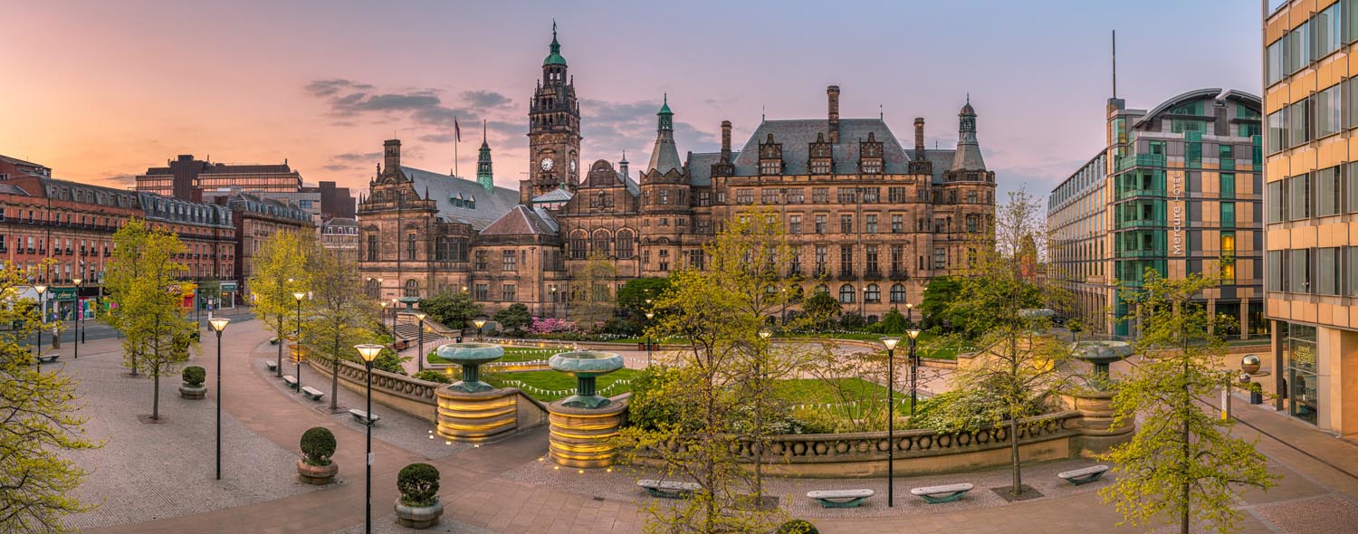 The Peace Gardens in Sheffield city centre; Panoramic view of a historic city square at sunset, featuring a large ornate building with towers and detailed architecture, likely a town hall. The square includes landscaped areas with trees, benches, and tiered fountains. Modern buildings border the scene, and the sky glows with warm orange and pink tones.