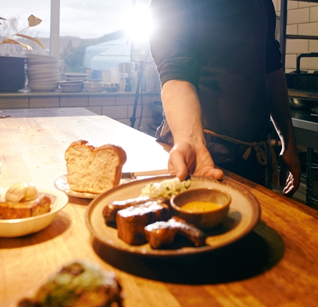A smiling man, in an apron, is placing a plate of food down on a table.