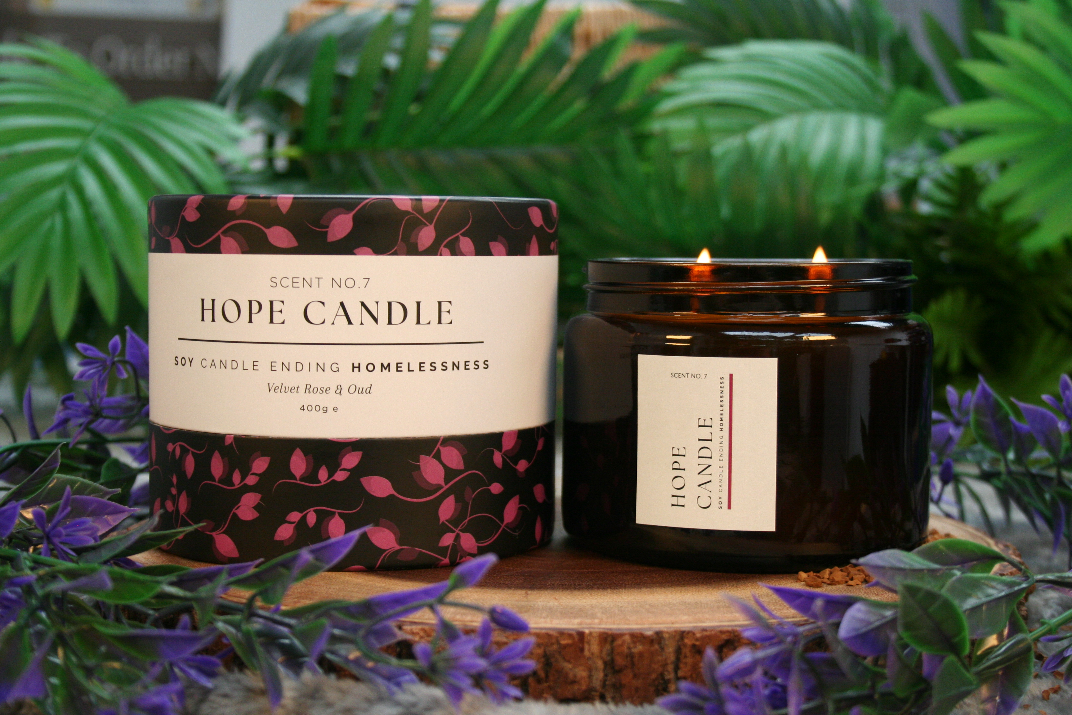 Hope Candle Scent No.7 in Velvet Rose & Oud fragrance, featuring a black and pink floral-patterned box and a lit amber glass jar candle with two wicks, displayed on a wooden base surrounded by green leaves and purple flowers.