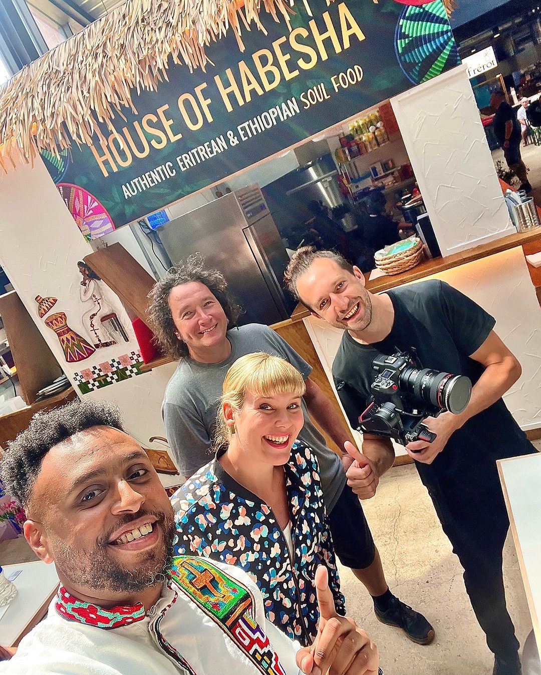 Group of people standing inside a food stall named ‘House of Habesha,’ which has a sign reading ‘Authentic Eritrean & Ethiopian Soul Food.’ The stall features cultural artwork on the walls, and one person is holding a professional camera