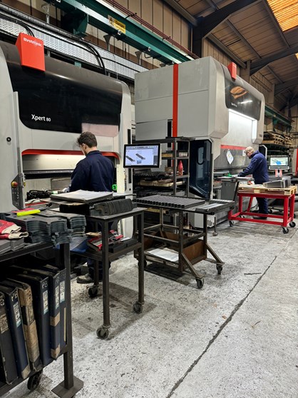 Industrial workshop with two large press brake machines labeled ‘Bystronic Xpert 80.’ Workstations with metal parts, tools, and a computer monitor displaying technical diagrams are positioned in front of the machines. Overhead beams and shelving with materials are visible in the background.