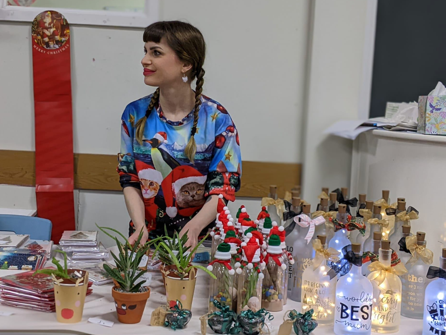Indoor craft stall with a festive display. The table is decorated with small potted plants, handmade ornaments, and decorative bottles with lights and ribbons. Some bottles have messages like “Best Mum” and “Christmas Magic.” There are also Christmas cards stacked on one side and miniature Santa figurines arranged in glass containers. The person behind the table is wearing a colourful Christmas-themed sweater featuring Santa and animals.