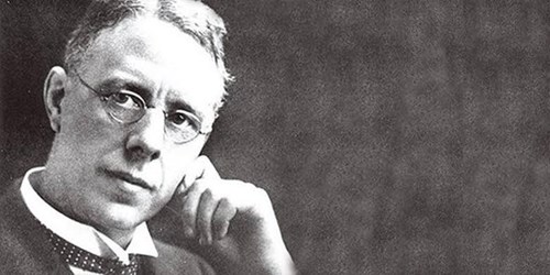Black-and-white photograph of a person wearing a formal suit with a collared shirt and tie, seated and resting one arm on a surface while the other hand is positioned near the chin. The background is plain and dark, creating a classic portrait style.