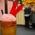 A closeup of a colourful cocktail, in the background are two people standing at the bar at the Parrot Club.