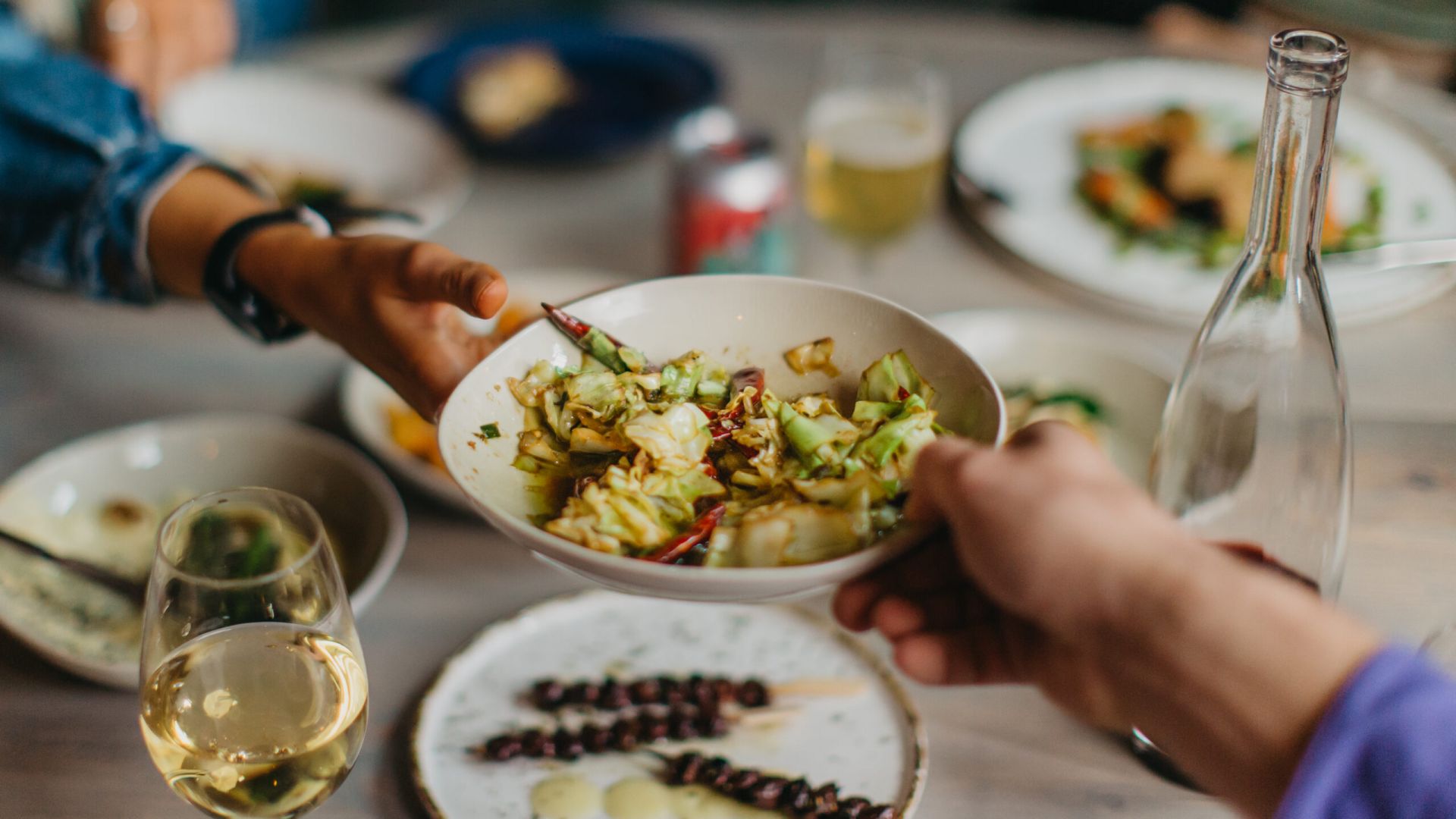 A close-up of a dining table with several dishes and drinks. In the foreground, two hands are passing a white bowl filled with a vegetable dish that includes chopped greens and red chili peppers. Surrounding the bowl are plates with various foods, a glass of white wine, a clear glass bottle, and a can of soda. The table setting suggests a shared meal in a casual, social atmosphere.