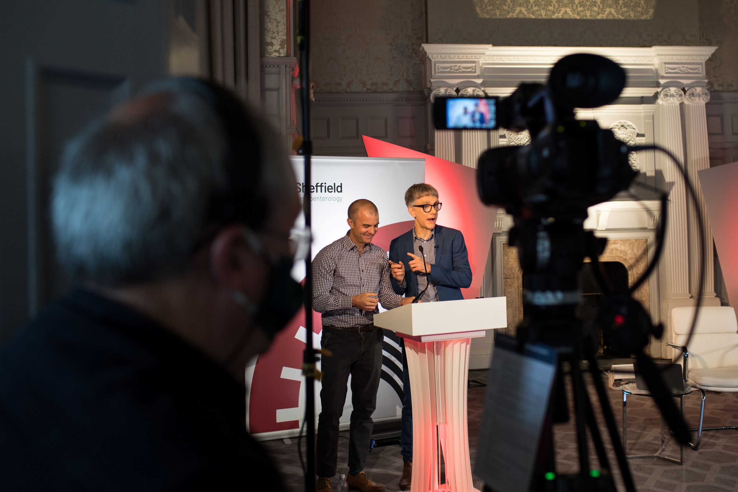 Two presenters standing behind a white podium on a stage, speaking into microphones during an event. A professional video camera on a tripod is positioned in the foreground, filming the scene. The backdrop includes event branding with the word ‘Sheffield’ partially visible, and the setting features ornate wall panelling and decorative architecture.