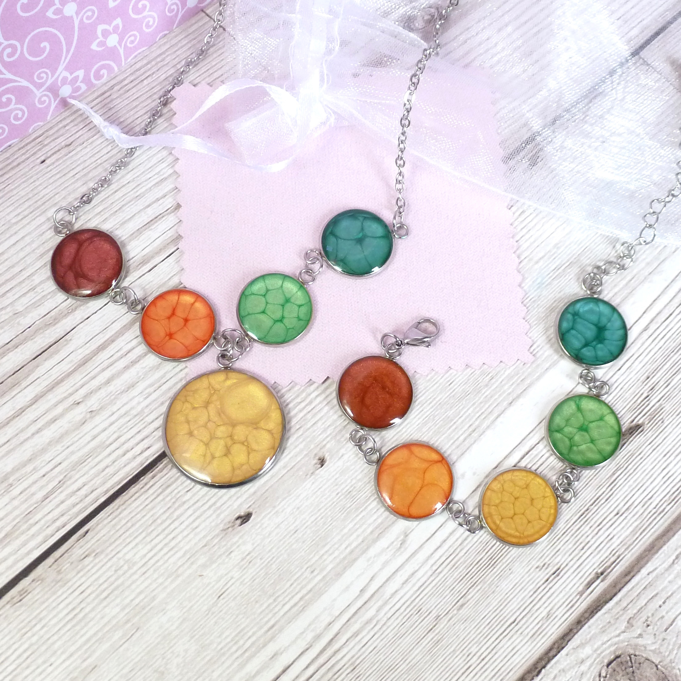 Handmade necklace and bracelet set featuring round resin bezels in vibrant shades of yellow, orange, green, and red with crackle texture, displayed on a light wooden surface with pink and white packaging.
