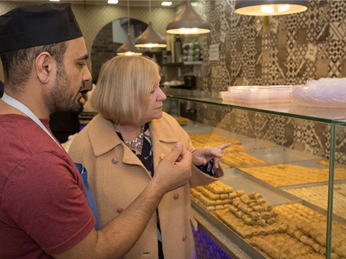 Two people stand in front of a glass display case filled with neatly arranged pastries. One wears a red shirt and black cap, the other a beige coat. They appear to be discussing the selection. The background features patterned tiles and hanging lights, suggesting a bakery or pastry shop setting.