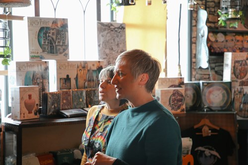 Two people standing in a warmly lit store filled with Harry Potter merchandise, including puzzles and games on shelves. The space has a cosy atmosphere with decorative plants and hanging lights.