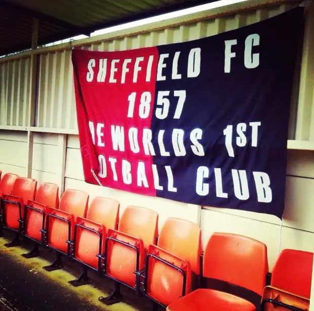 A banner hanging on a corrugated wall behind a row of red stadium seats. The banner is split into red and black halves and reads in bold white letters: ‘Sheffield FC 1857 The World’s 1st Football Club.’