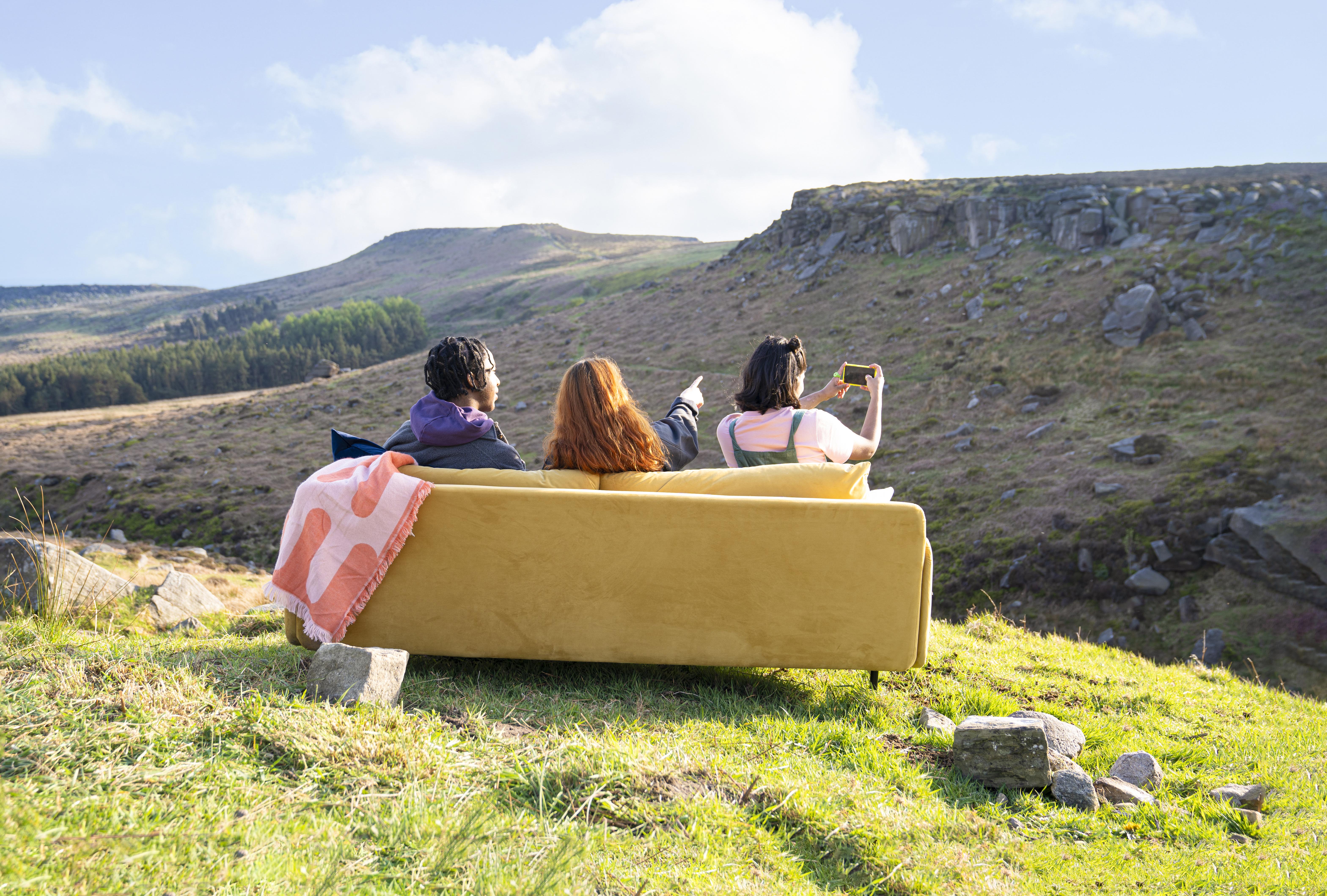 Three people sitting on a yellow couch placed outdoors on a grassy hillside, overlooking a rugged landscape with rocky terrain and distant hills under a bright blue sky with scattered clouds. One person is pointing toward the horizon while another holds up a phone, possibly taking a photo.