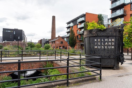 Outdoor view of Kelham Island Museum in Sheffield. A large industrial metal container with “Kelham Island Museum” written on it stands in the foreground near railings and a water channel. Surrounding the area are modern apartment buildings, a tall brick chimney, and greenery under a cloudy sky.