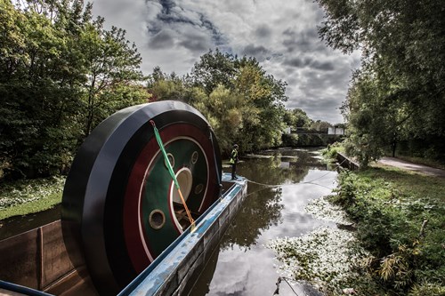 Large circular sculpture with green, red, and black rings being transported on a flat barge along a narrow canal, surrounded by dense trees and vegetation under a cloudy sky.