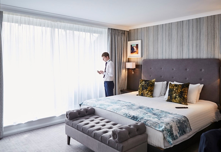 Elegant luxury bedroom featuring plush cushions on a king-sized bed, soft ambient lighting, and a large window with sheer curtains letting in natural light.