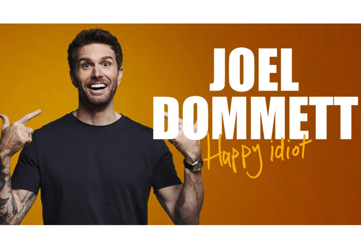 Joel Dommett is smiling and pointing at himself. Next to him it says 'Joel Dommett - Happy Idiot'.