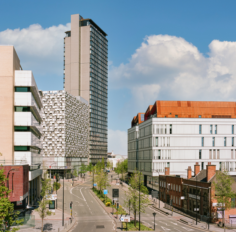 Urban street scene with modern architecture under a bright blue sky with scattered clouds. The view shows a wide road lined with trees and streetlights, flanked by contemporary buildings. Notable structures include a tall glass and concrete tower, a building with a distinctive patterned façade, and another with a rust-colored roof section. The street appears quiet with minimal traffic and clear pedestrian pathways.