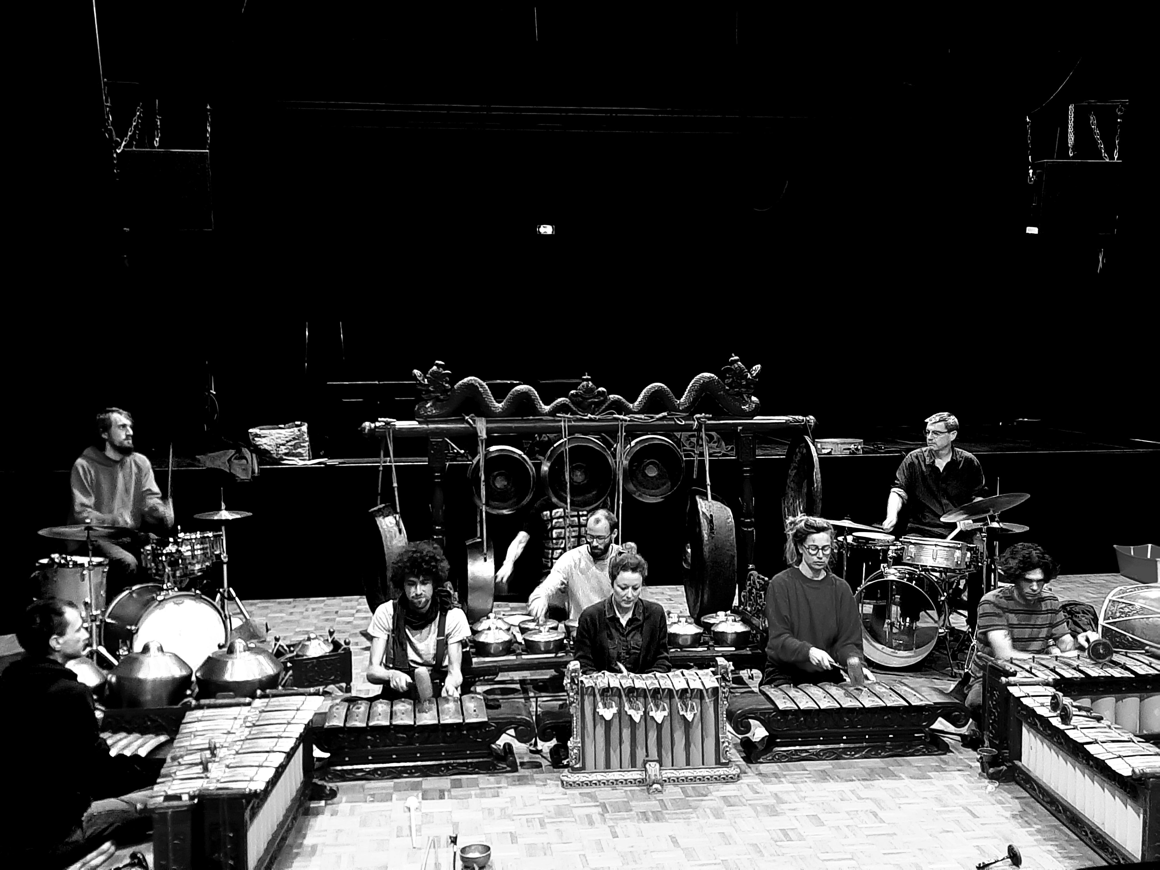 A black-and-white photo of a music ensemble performing on stage with traditional and modern instruments. The setup includes large hanging gongs in ornate frames at the center, surrounded by metallophones and xylophones in the foreground. Drum kits are positioned on both sides, and several musicians are seated or standing behind their instruments. The stage is dimly lit, emphasising the instruments and performers.