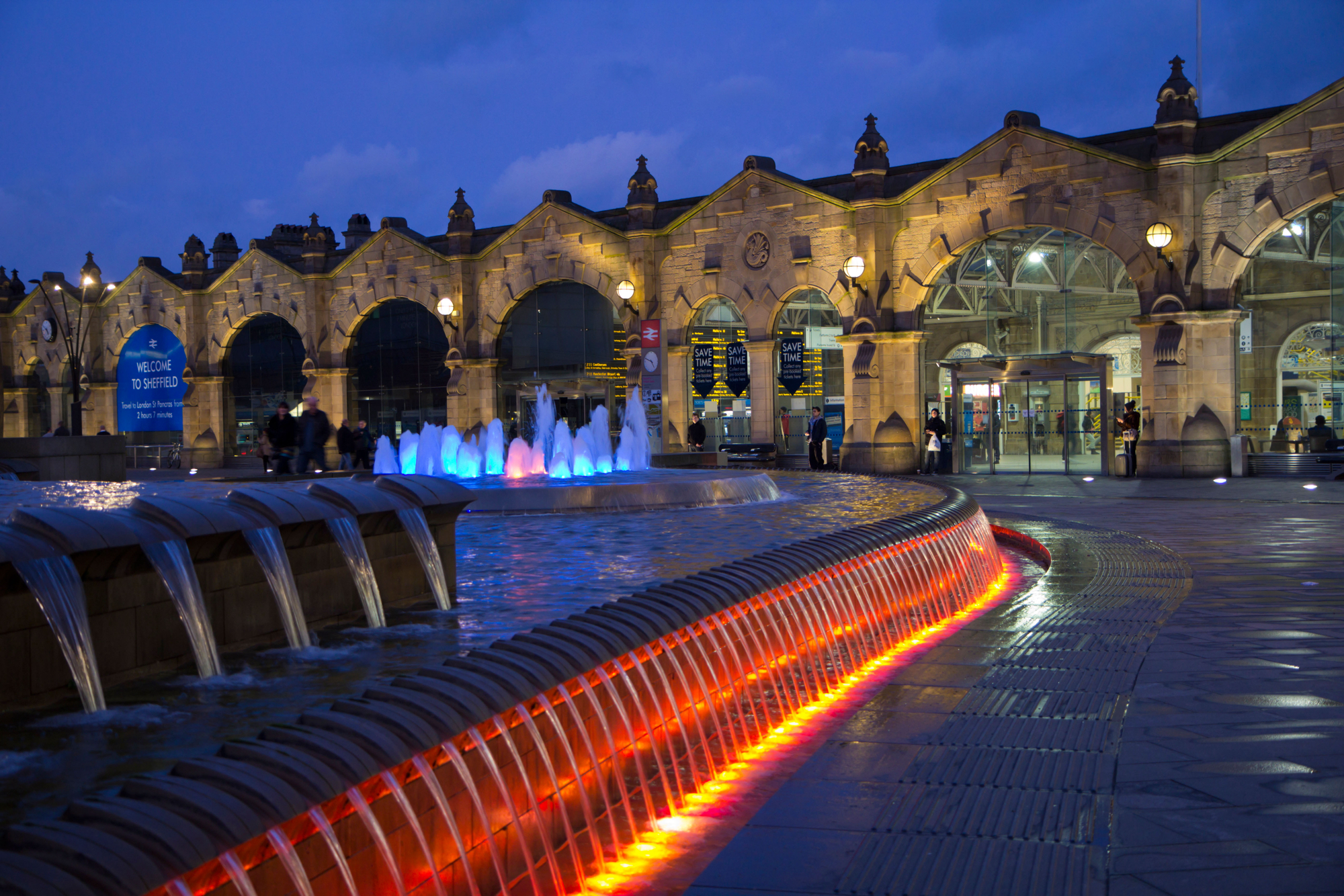 Night view of Sheffield railway station with its historic stone façade illuminated by warm lights. In the foreground, a curved water feature with cascading fountains is lit in vibrant colors, including red, orange, and blue. The station entrance and signage are visible in the background under a deep blue evening sky.