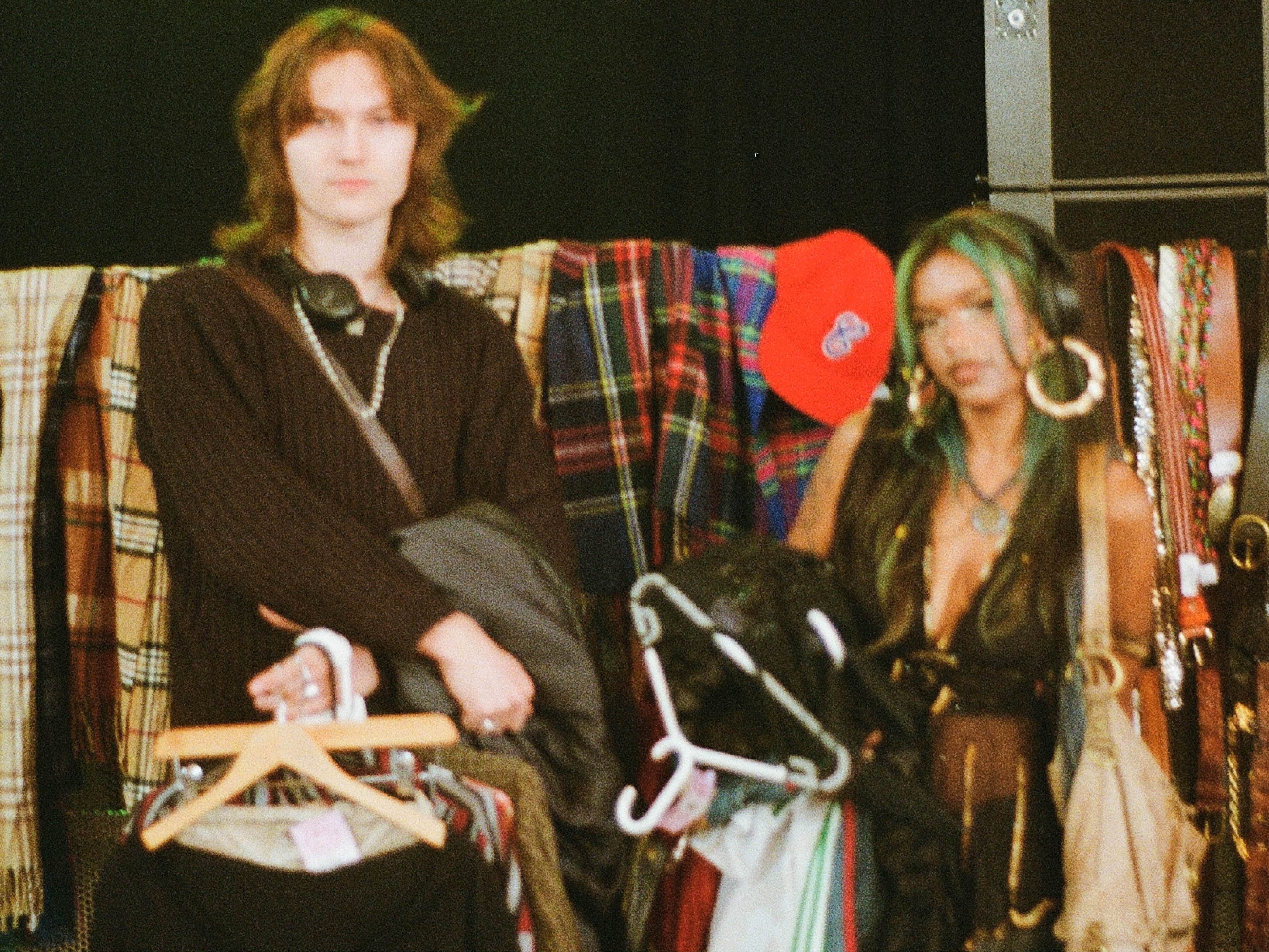 Two people are standing in front of a display of colorful plaid and patterned scarves or fabrics. One person is holding a hanger with multiple clothing items, while the other is carrying several garments draped over hangers. Behind them, a bright red baseball cap is visible among the fabrics. The setting appears to be a vintage or second-hand clothing market or store.