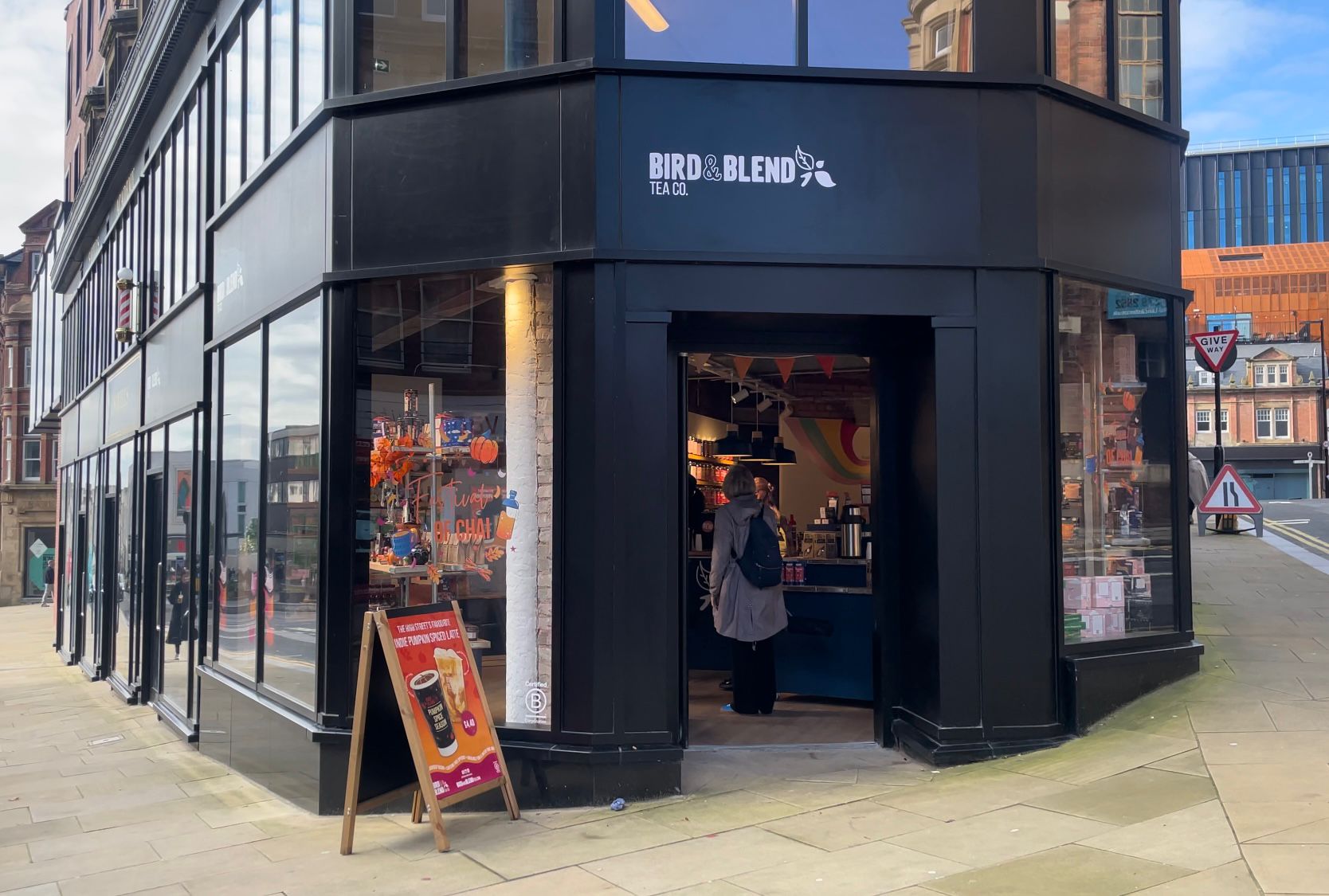 Exterior view of a corner tea shop called “Bird & Blend Tea Co.” with large glass windows and a black storefront. The entrance is open, revealing a colorful interior with hanging decorations and a counter. A sandwich board outside displays promotional drinks, and the shop is located on a paved street with buildings and traffic signs visible in the background.