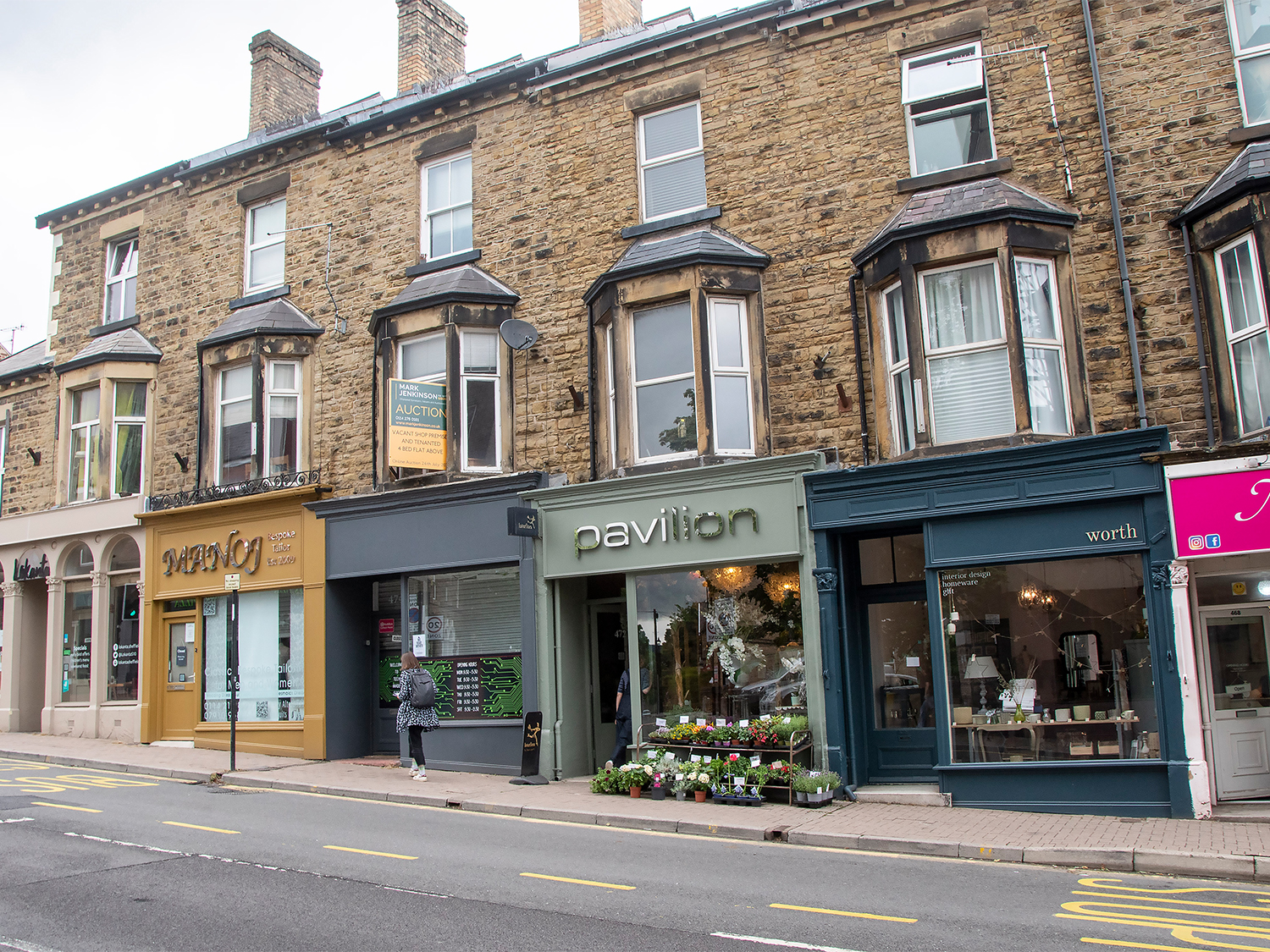 Row of stone-built Victorian-style buildings with ground-floor shops along a street. Visible storefronts include “Valley Insurance Services” with a mustard-yellow facade, “Pavilion” with green signage and flower display outside, and “Worth” with a dark blue front showcasing homeware items. A person stands near the insurance shop, and a “For Auction” sign is displayed in an upper window.