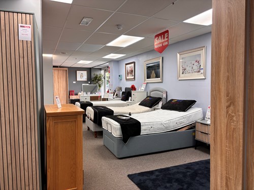 Interior of a bedroom showroom featuring two adjustable beds with white mattresses and black throws. The walls are decorated with framed artwork, and there are bedside tables, a chest of drawers, and a wooden cabinet in the foreground. A red sign hanging from the ceiling reads ‘SALE NOW ON.’ Bright overhead lighting illuminates the space.