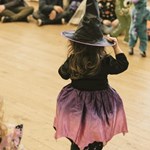 Child in a witch costume with a black pointed hat and a purple-to-pink ombre dress, standing on a wooden floor facing away. Background shows other children and adults, some in costumes, at an indoor event or party.