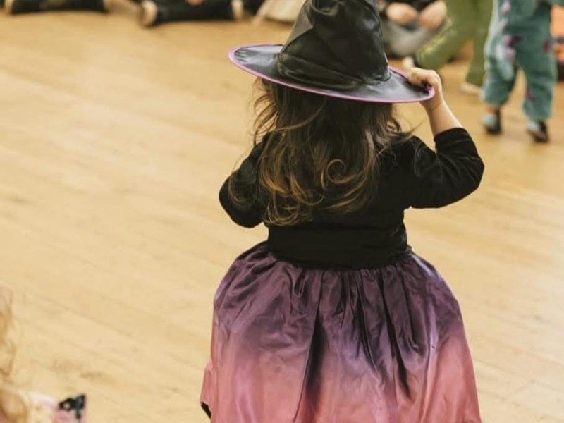 Child in a witch costume with a black pointed hat and a purple-to-pink ombre dress, standing on a wooden floor facing away. Background shows other children and adults, some in costumes, at an indoor event or party.