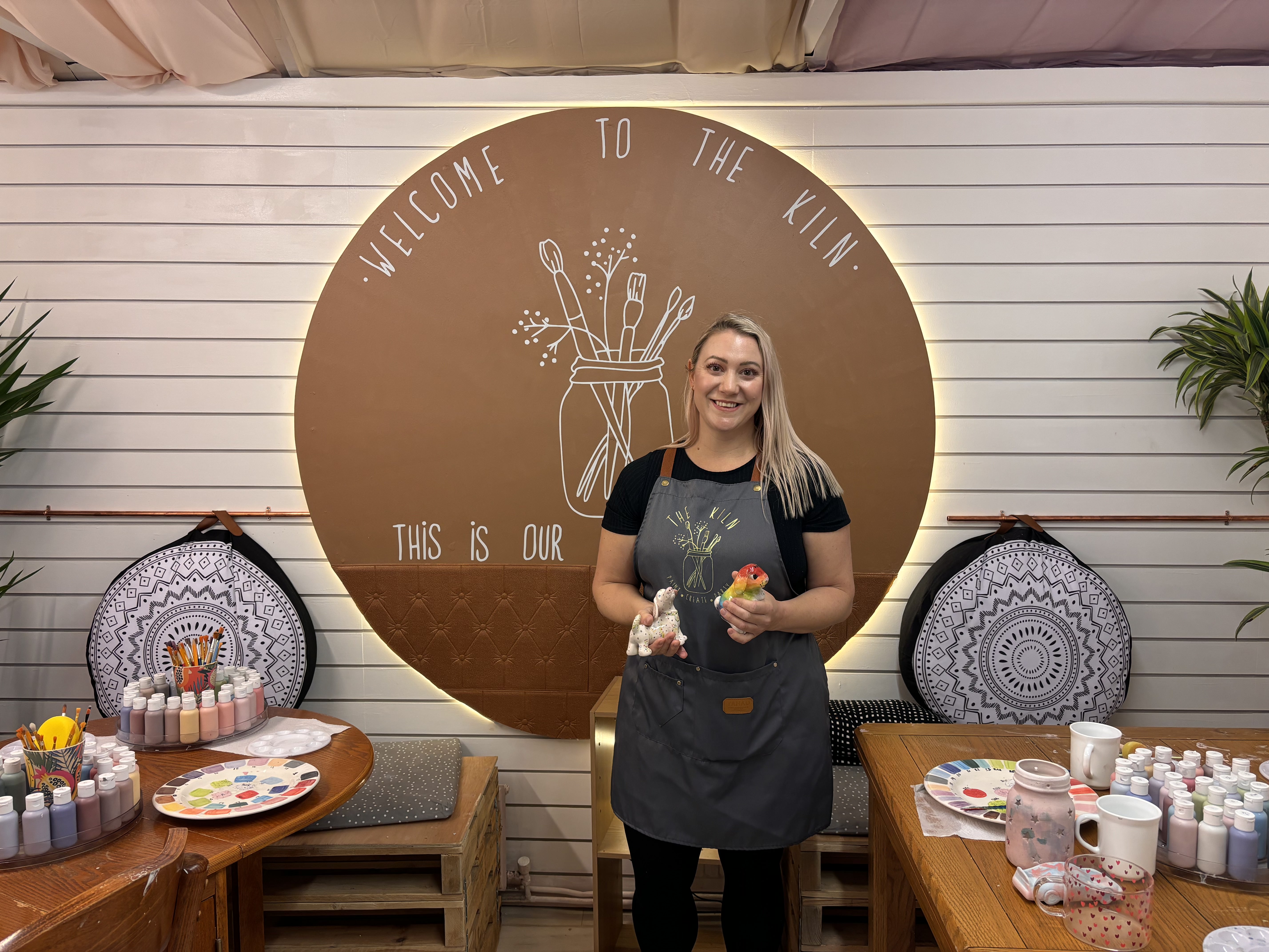 Person wearing a gray apron standing in front of a circular sign that reads ‘Welcome to the Kiln’ and ‘This is our…’ with an illustration of paintbrushes. The setting includes wooden tables with paint bottles, ceramic items, and color palettes arranged for a pottery painting activity. Decorative cushions and potted plants are visible in the background.