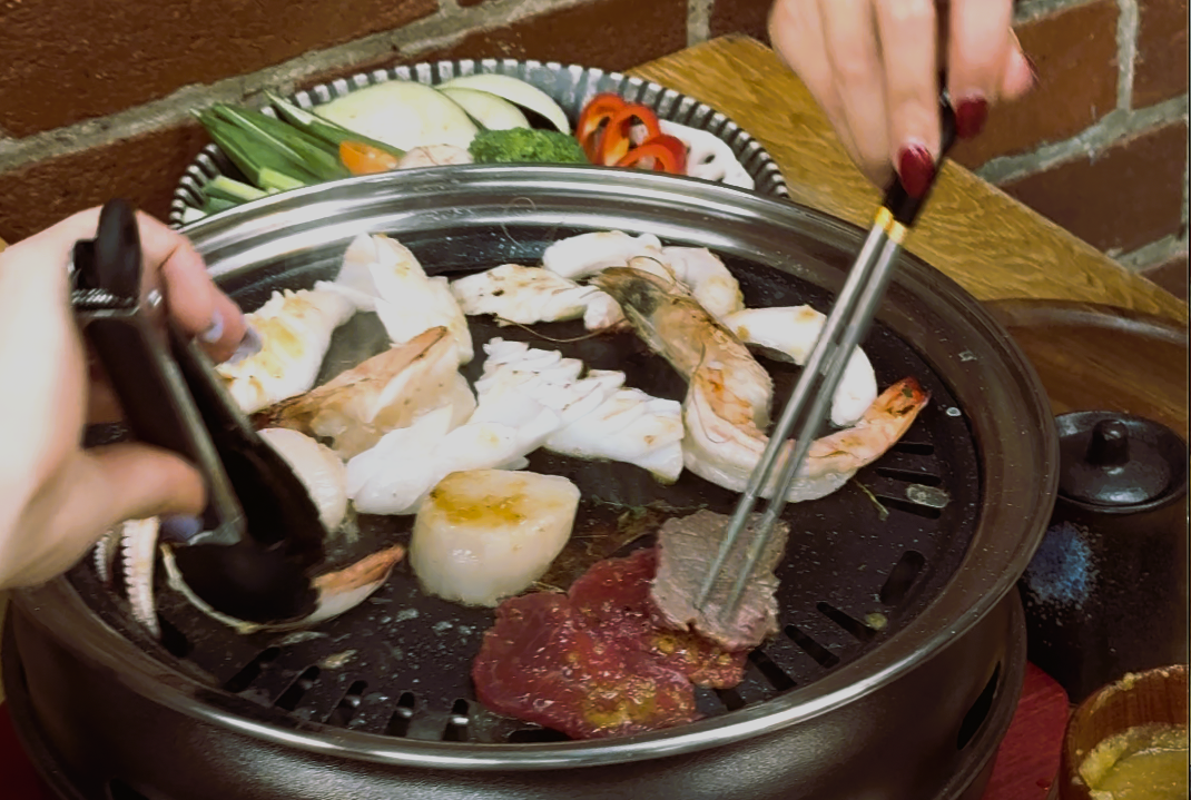 Close-up of a tabletop grill with various seafood and meat pieces cooking, including shrimp, scallops, and beef slices. Two hands are using tongs and chopsticks to turn the food. In the background, a plate of fresh vegetables such as zucchini, bell peppers, and green onions is visible.