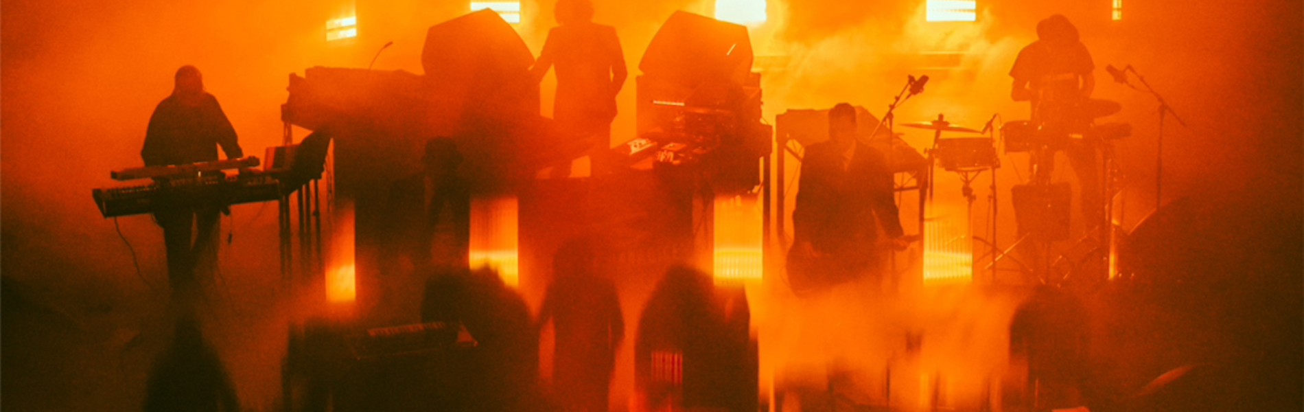 Silhouetted musicians perform on stage surrounded by dense orange fog and dramatic vertical light beams. The scene is bathed in warm, glowing tones, creating an abstract, atmospheric effect with blurred reflections on the floor.