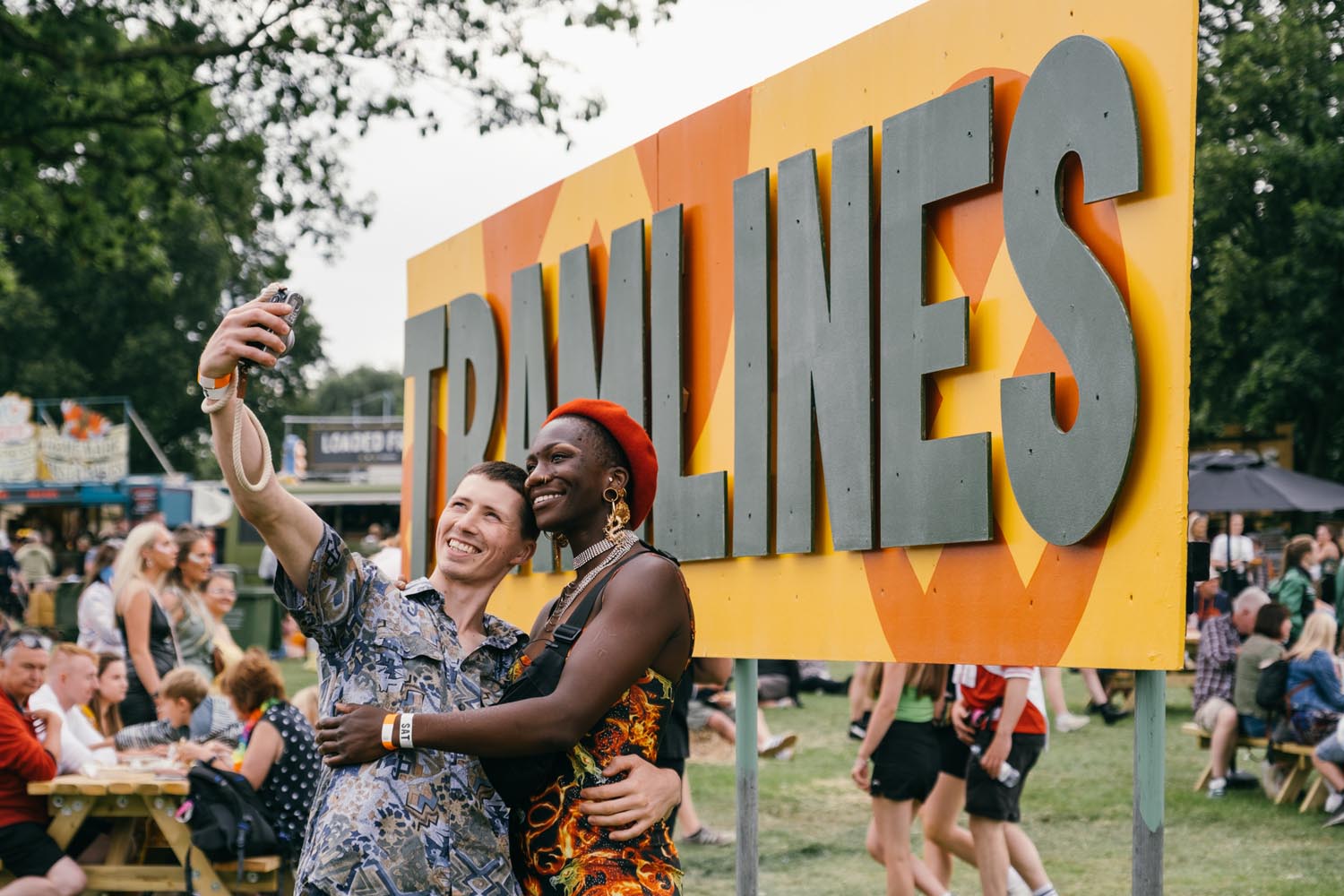 Two people taking a selfie in front of a large yellow and orange sign that reads “TRAMLINES” in bold gray letters. The scene is set at an outdoor festival with grass underfoot, picnic tables, and groups of people sitting and walking in the background. Food stalls and trees are visible further back, creating a lively and colorful atmosphere.