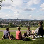 Four people are sat on the grass at Meersbrook Park in the sun. In the distance you can see Sheffield city centre.