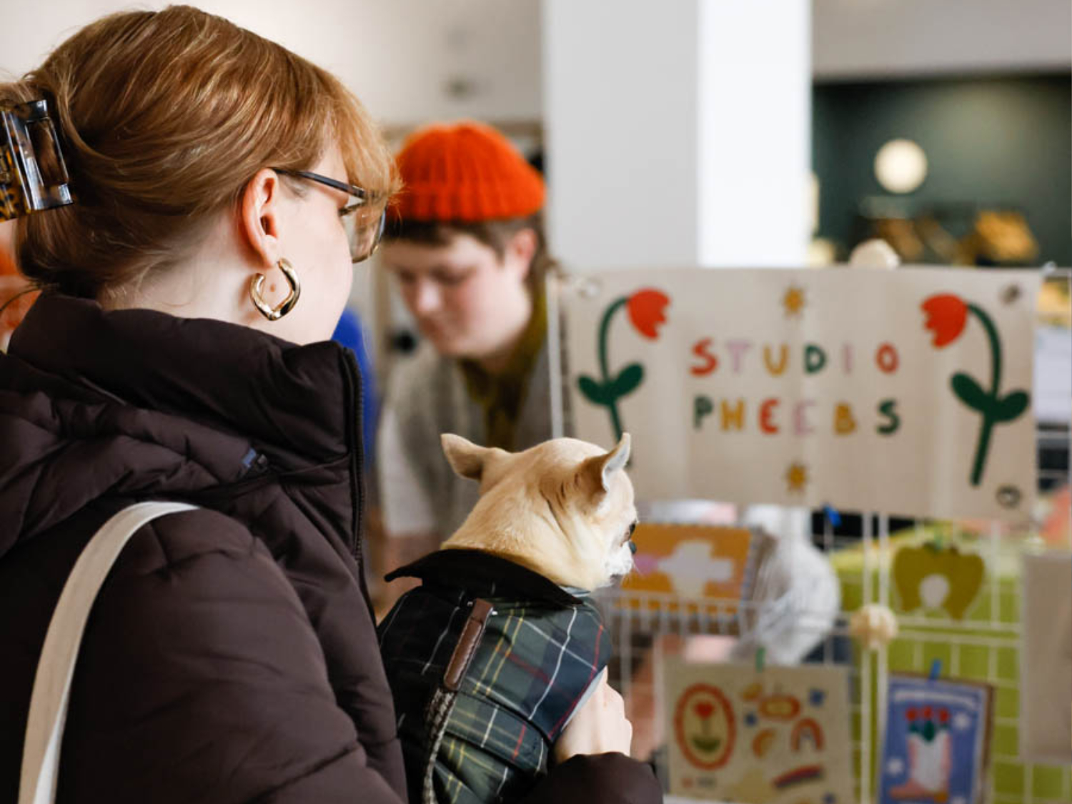 A person wearing glasses and a dark jacket holds a small dog in a plaid coat in front of a booth labeled "STUDIO PHEEBS," decorated with colorful flowers. Another person in an orange beanie stands behind the booth, which displays various art or craft items, suggesting a lively market or fair setting.