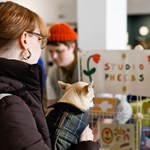 A person wearing glasses and a dark jacket holds a small dog in a plaid coat in front of a booth labeled "STUDIO PHEEBS," decorated with colorful flowers. Another person in an orange beanie stands behind the booth, which displays various art or craft items, suggesting a lively market or fair setting.